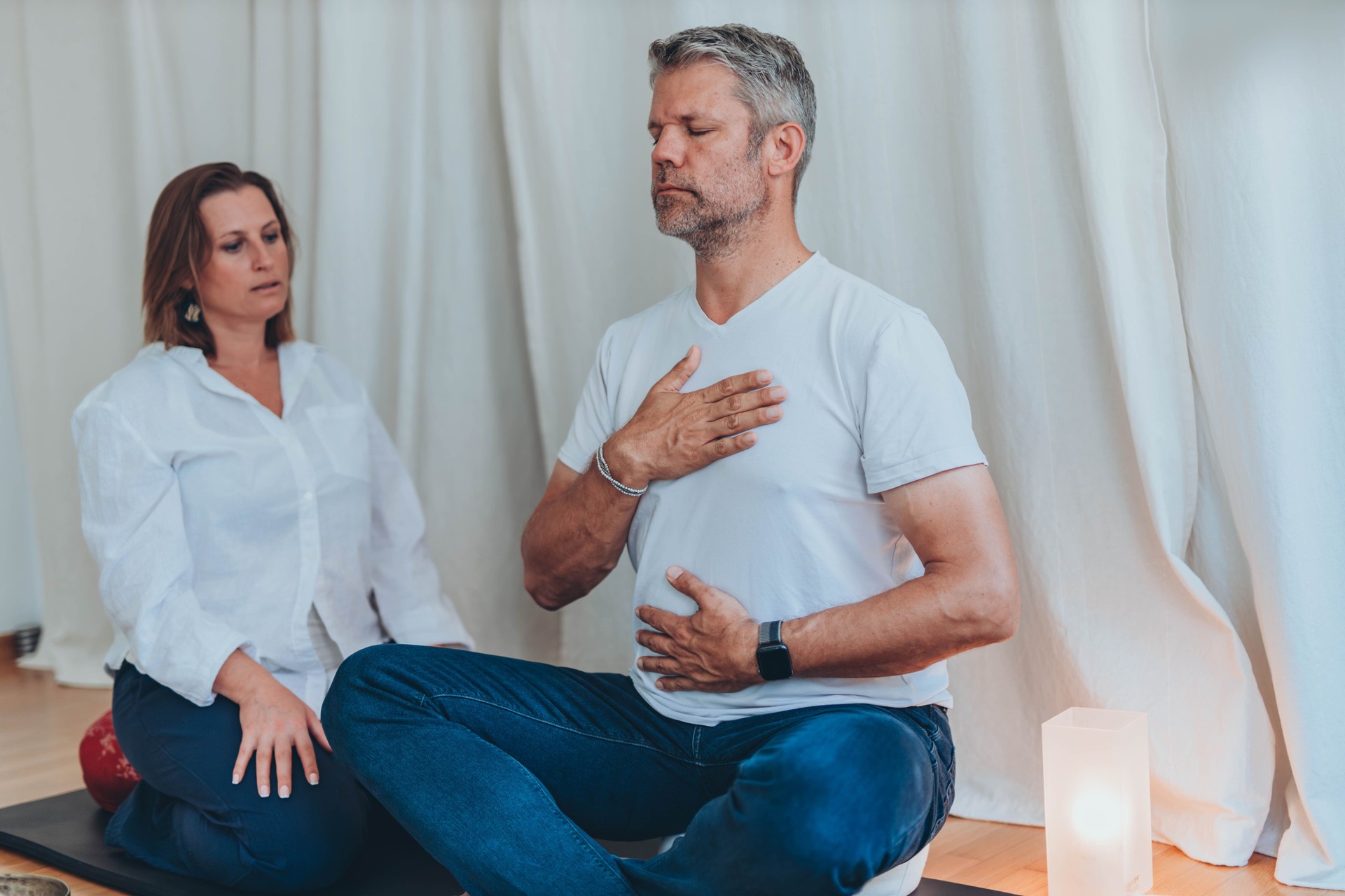 A man and a woman sitting in lotus position, the man wearing a blindfold while practicing breathing exercises with the woman's coaching